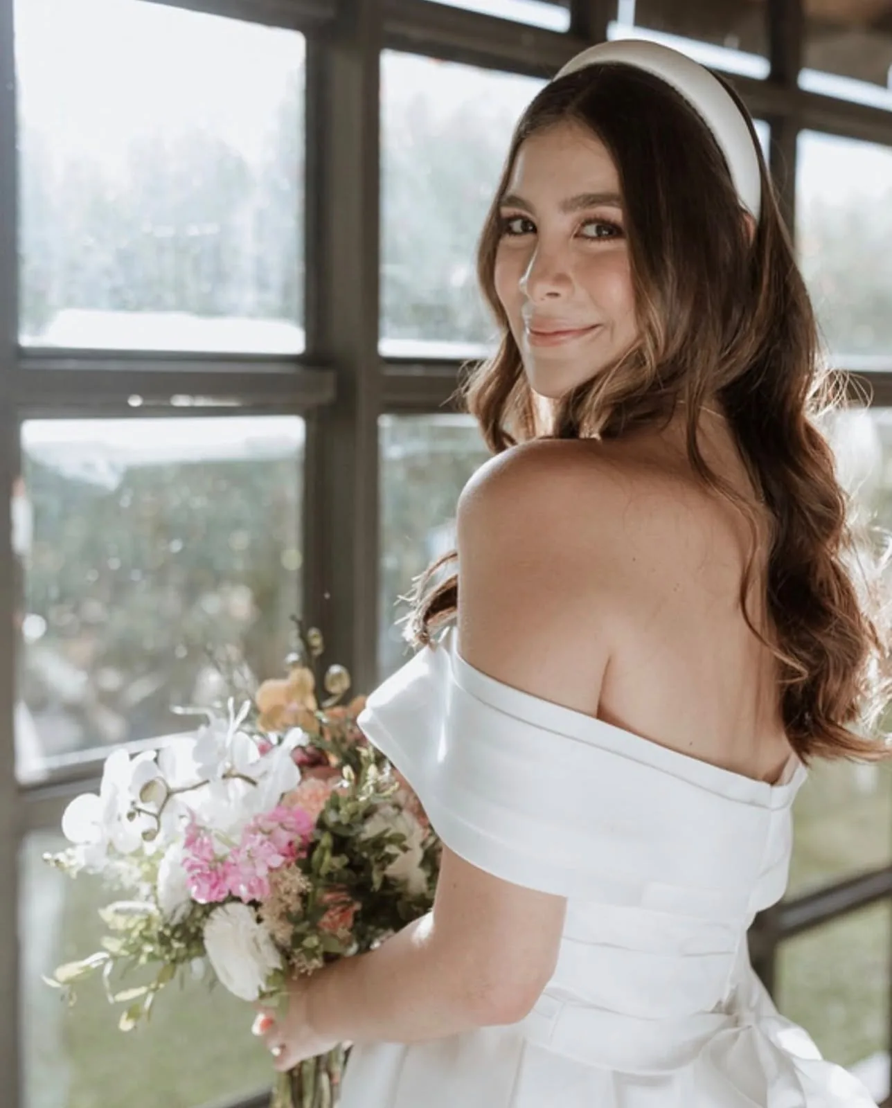 Bride looking over her shoulder holding a flower bouquet by a window