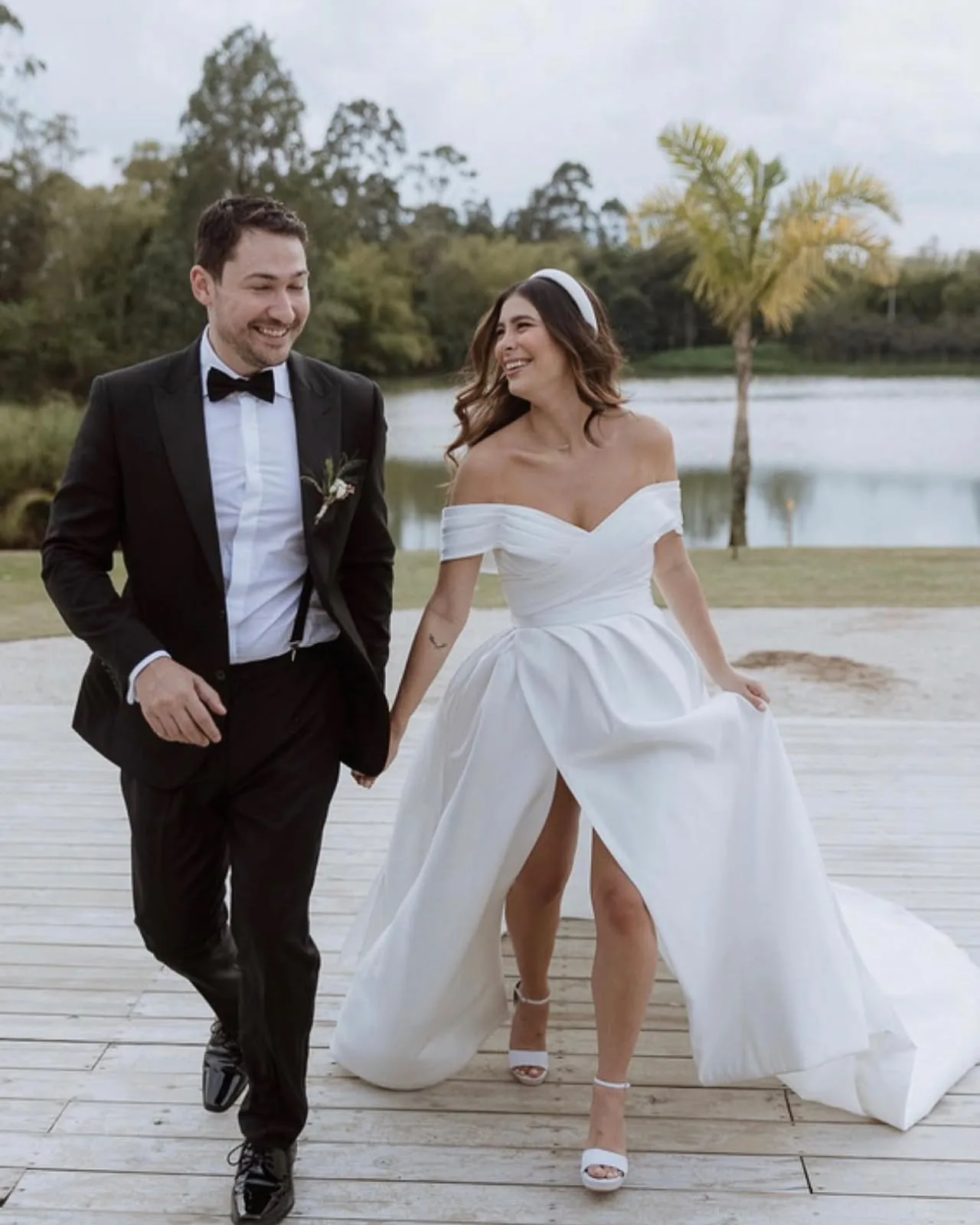 Newlyweds walking beside a lake at sunset, bride in a low-cut gown with a high slit