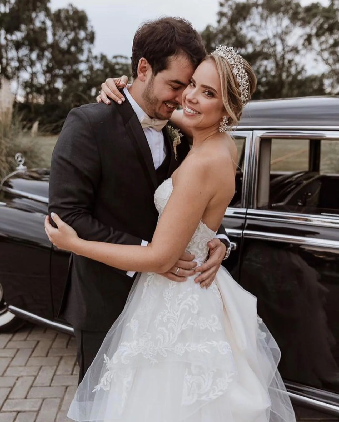Newlyweds embracing next to a classic car, bride wearing a crystal tiara and lace gown