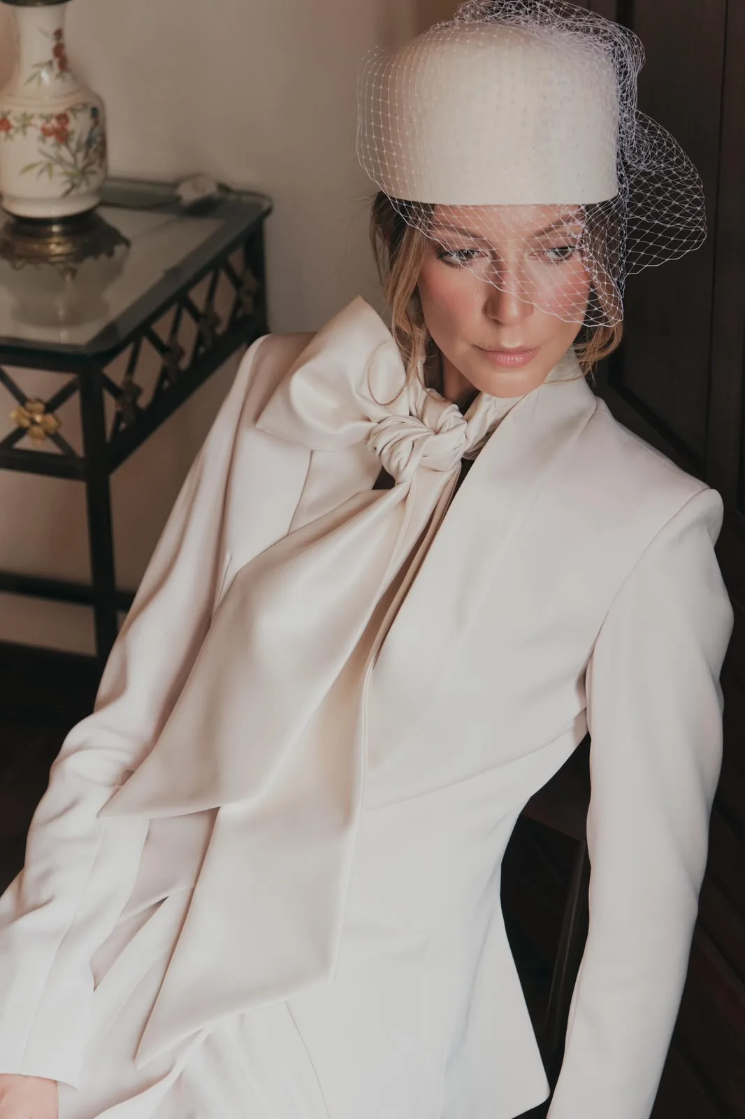 Model wearing a birdcage headpiece and a cream tailored suit with a silk bow